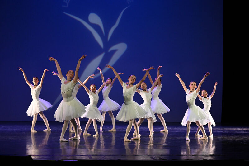 Group of Ananyan Ballet students in white tutus performing on a blue-lit stage with arms raised.