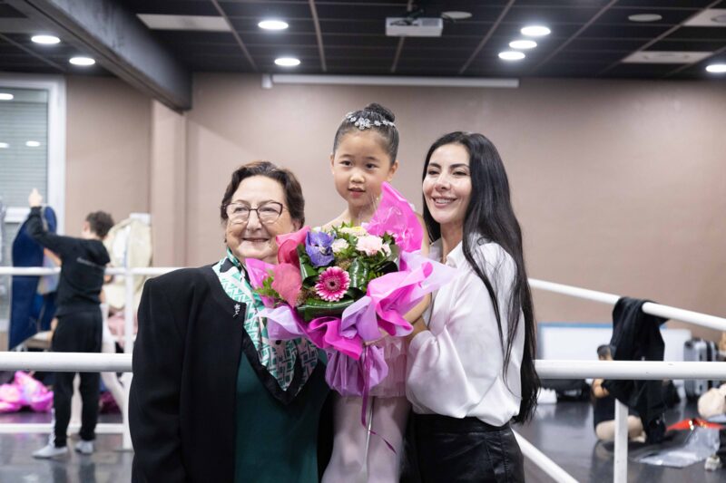 Victoria Ananyan backstage with a young student holding a bouquet of pink flowers and another adult.